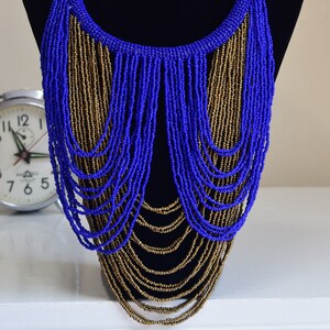 May include: A blue and gold beaded necklace with a cascading design. The necklace is displayed on a black mannequin bust.