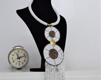 May include: A long white beaded necklace with two large white and colorful beaded circles and a gold accent. The necklace is displayed on a black mannequin.