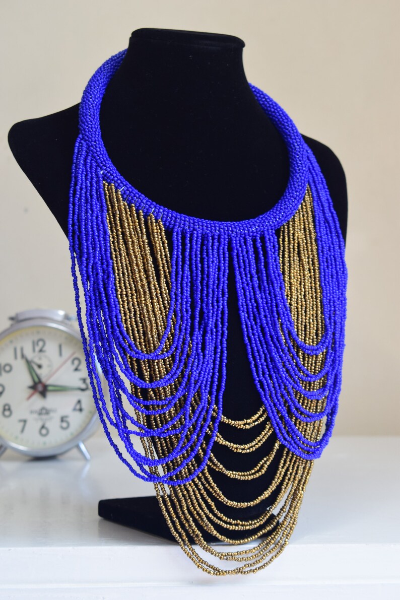 May include: A long, layered necklace made of blue and gold beads. The necklace is displayed on a black mannequin bust.