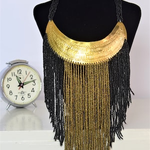 Könnte beinhalten: Eine goldene und schwarze Perlenkette mit einem großen, halbmondförmigen Anhänger. Der Anhänger hat eine strukturierte Oberfläche und ist aus Metall.