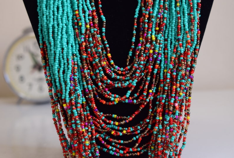 Puede incluir: Un collar de cuentas de m&uacute;ltiples hilos con cuentas turquesa, rojas, naranjas, amarillas y moradas. El collar est&aacute; en capas y cuelga en la parte delantera del cuello.