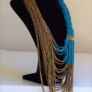 May include: A long, layered necklace with a braided turquoise section and gold accents. The necklace is made of small beads and is displayed on a black mannequin.