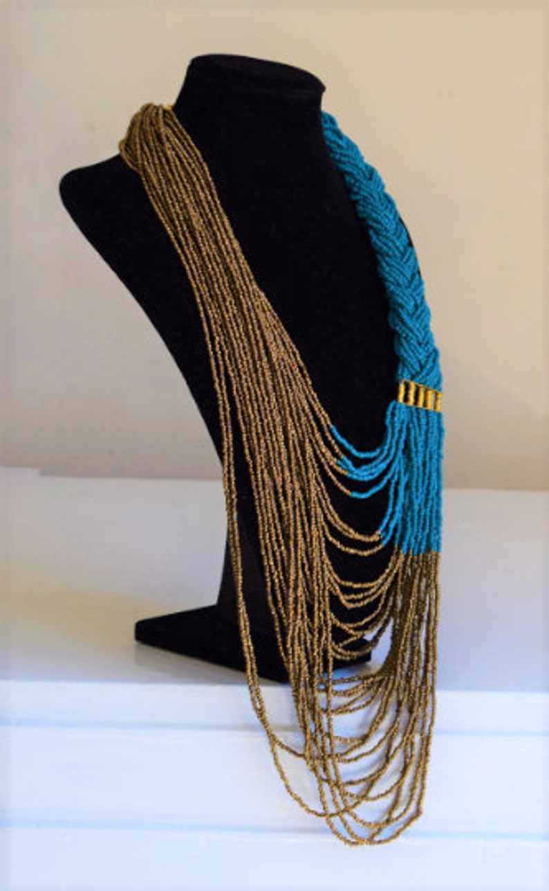 May include: A long, layered necklace with a braided turquoise section and gold accents. The necklace is made of small beads and is displayed on a black mannequin.