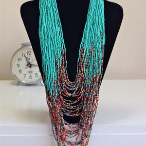 Puede incluir: Un collar largo de cuentas turquesa y rojo con m&uacute;ltiples hebras. El collar se muestra en un maniqu&iacute; negro.