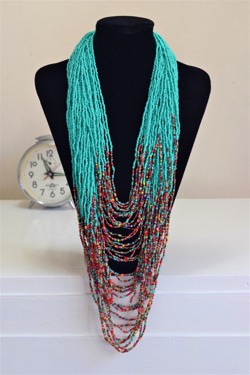 Puede incluir: Un collar largo de cuentas turquesa y rojo con m&uacute;ltiples hebras. El collar se muestra en un maniqu&iacute; negro.