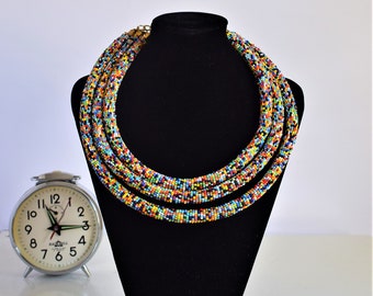 May include: Multicolored beaded necklace on a black velvet display stand. The necklace is made of small, colorful beads and is layered in a circular pattern.