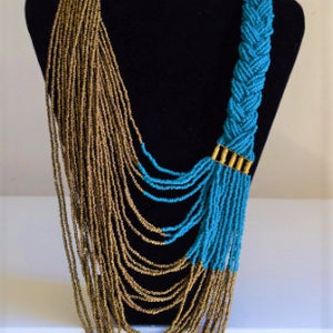 Op de afbeelding: Een lange, gelaagde ketting met een gevlochten gedeelte van turquoise blauwe kralen en een waterval van gouden kralen.