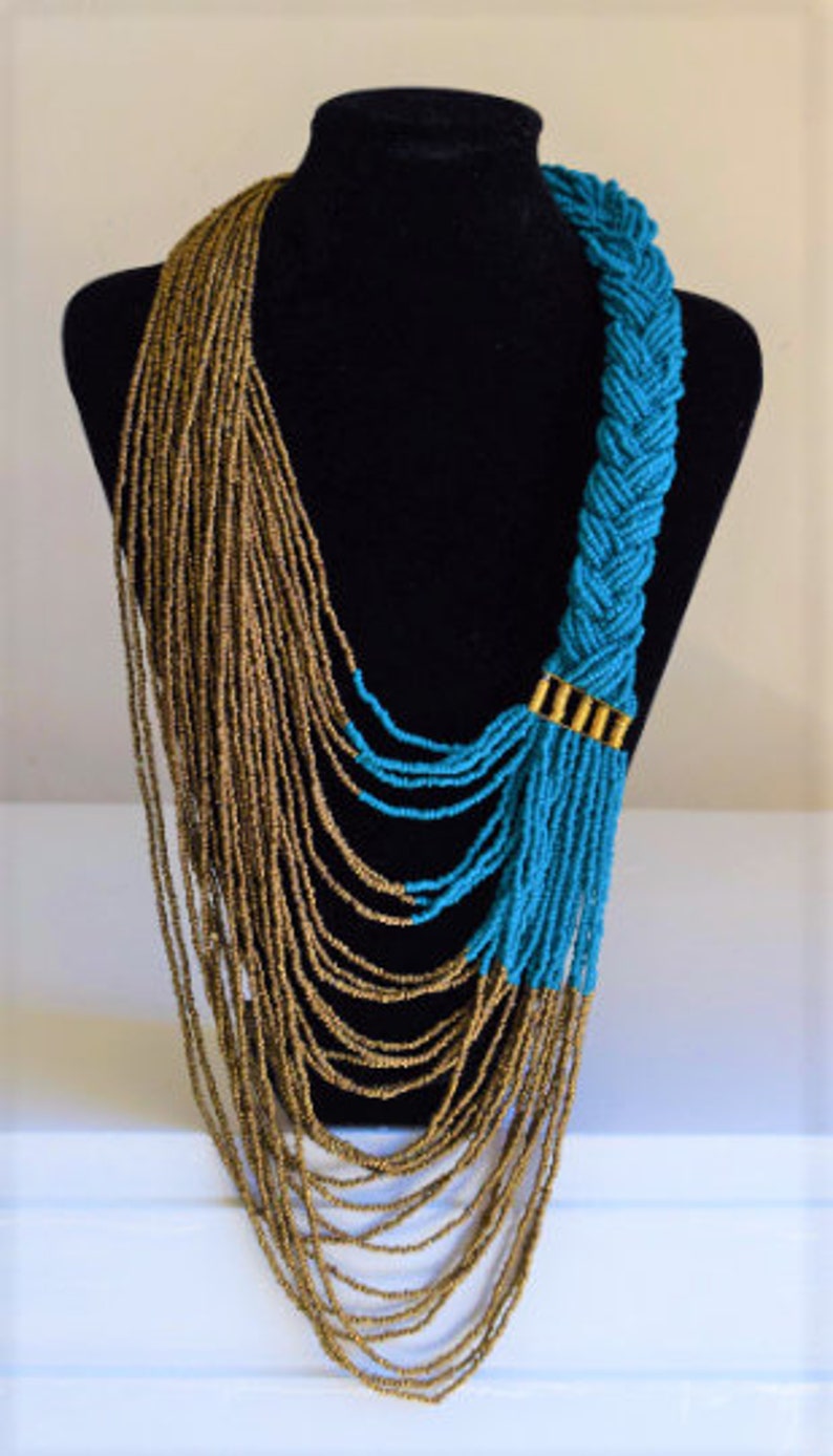Op de afbeelding: Een lange, gelaagde ketting met een gevlochten gedeelte van turquoise blauwe kralen en een waterval van gouden kralen.