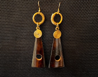 May include: A pair of brown wooden earrings with gold wire accents. The earrings are shaped like a triangle with a round hole in the center. The earrings are hanging from a gold wire loop.