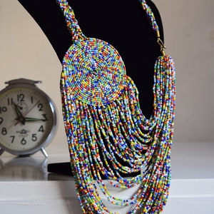 May include: A colorful beaded necklace with a large circular pendant. The necklace is made of small, multi-colored beads and has a long, cascading design.