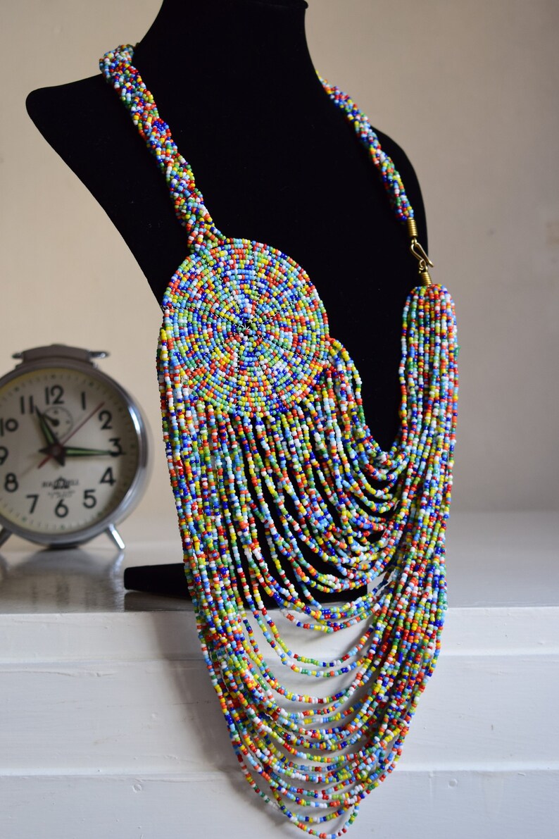 May include: A colorful beaded necklace with a large circular pendant. The necklace is made of small, multi-colored beads and has a long, cascading design.