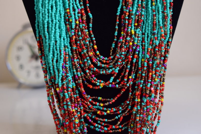 Puede incluir: Un collar de cuentas de m&uacute;ltiples hilos con cuentas turquesa, rojas, naranjas, amarillas y moradas. El collar est&aacute; en capas y cuelga en la parte delantera del cuello.