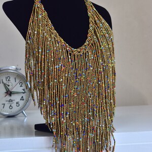 May include: A long gold beaded necklace with colourful beads woven throughout. The necklace is on a black mannequin and has a fringe design.