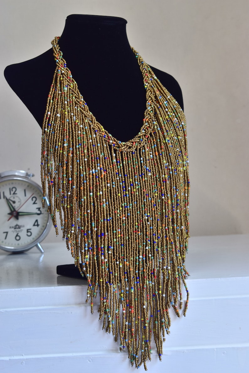 May include: A long gold beaded necklace with colourful beads woven throughout. The necklace is on a black mannequin and has a fringe design.