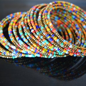 Puede incluir: Un conjunto de pulseras de cuentas de colores con detalles dorados. Las pulseras están hechas de pequeñas cuentas redondas de varios colores, incluyendo rojo, naranja, amarillo, verde, azul y morado. Las pulseras están dispuestas en espiral, con los detalles dorados visibles en todo el conjunto.