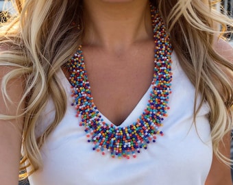 May include: A multi-strand statement necklace featuring a dense arrangement of small, vibrant seed beads in shades of red, blue, yellow, green, and white. The beads are strung together to create a textured, layered collar effect around the neck.