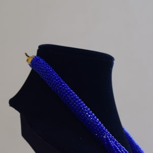 May include: A blue beaded necklace displayed on a black velvet stand. The necklace is made from small, round beads that are strung together in a long strand.