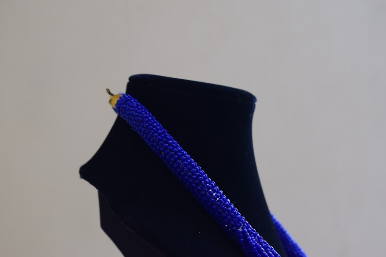 May include: A blue beaded necklace displayed on a black velvet stand. The necklace is made from small, round beads that are strung together in a long strand.