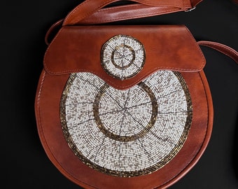 May include: A brown leather crossbody bag with a circular beaded design in white and gold. The bag has a flap closure and a long adjustable strap.