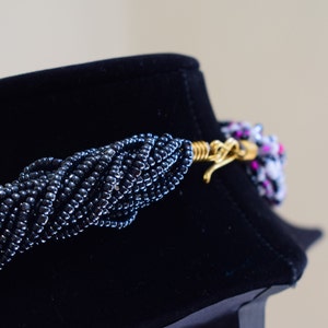 May include: A black beaded necklace with a gold clasp. The necklace is on a black velvet display stand.