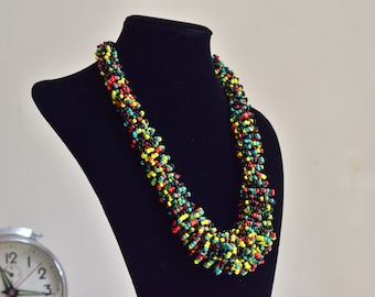 May include: A beaded necklace with a multi-colored design. The necklace is made of black, red, yellow, and green beads. The necklace is displayed on a black mannequin.