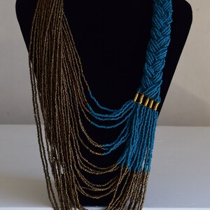 May include: A long, layered necklace with a braided section. The necklace is made of small, gold and teal beads. The braided section is teal and has a gold accent.