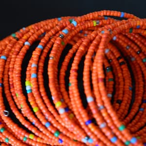 May include: A close-up of a stack of orange beaded bracelets. The bracelets are made of small, round beads with a few colorful beads interspersed throughout.