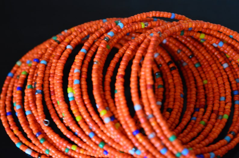May include: A close-up of a stack of orange beaded bracelets. The bracelets are made of small, round beads with a few colorful beads interspersed throughout.