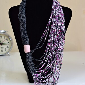 May include: A long, layered necklace made of black, white, and pink beads. The necklace is draped over a black mannequin bust. The necklace has a pink bead accent.