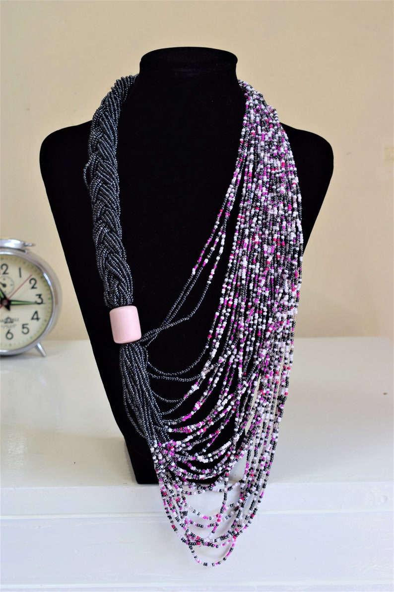 May include: A long, layered necklace made of black, white, and pink beads. The necklace is draped over a black mannequin bust. The necklace has a pink bead accent.