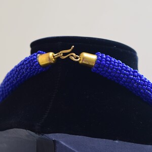 May include: A close-up of a blue beaded necklace with a gold clasp. The necklace is on a black velvet display stand.