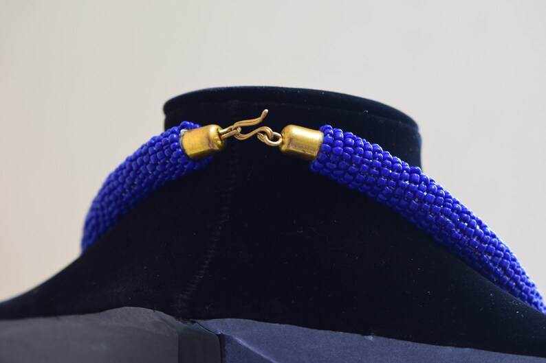 May include: A close-up of a blue beaded necklace with a gold clasp. The necklace is on a black velvet display stand.