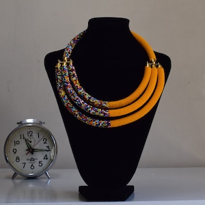 May include: A multi-strand beaded necklace displayed on a black mannequin. The necklace features three strands of bright yellow beads, transitioning to a colorful beaded pattern. Gold-colored clasps secure the necklace. A vintage alarm clock sits to the left.