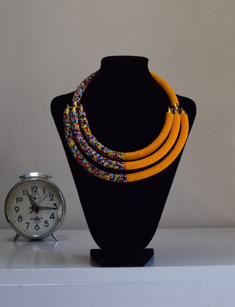 May include: A multi-strand beaded necklace displayed on a black mannequin. The necklace features three strands of bright yellow beads, transitioning to a colorful beaded pattern. Gold-colored clasps secure the necklace. A vintage alarm clock sits to the left.