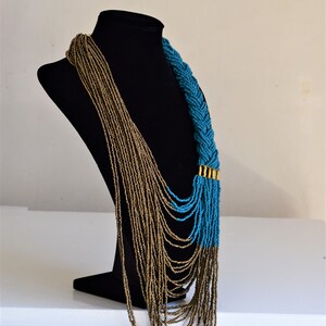 May include: A long, layered necklace with a braided section. The necklace is made of small, gold and turquoise beads. The necklace is displayed on a black mannequin.