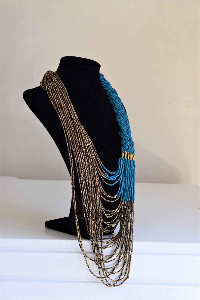May include: A long, layered necklace with a braided section. The necklace is made of small, gold and turquoise beads. The necklace is displayed on a black mannequin.