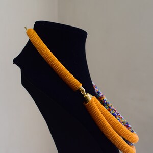 May include: A handmade necklace displayed on a black mannequin. The necklace features a vibrant orange beaded design with gold-colored clasps. A section of the necklace displays a colorful pattern of blue, red, and yellow beads.