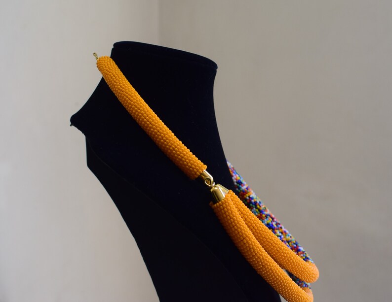 May include: A handmade necklace displayed on a black mannequin. The necklace features a vibrant orange beaded design with gold-colored clasps. A section of the necklace displays a colorful pattern of blue, red, and yellow beads.