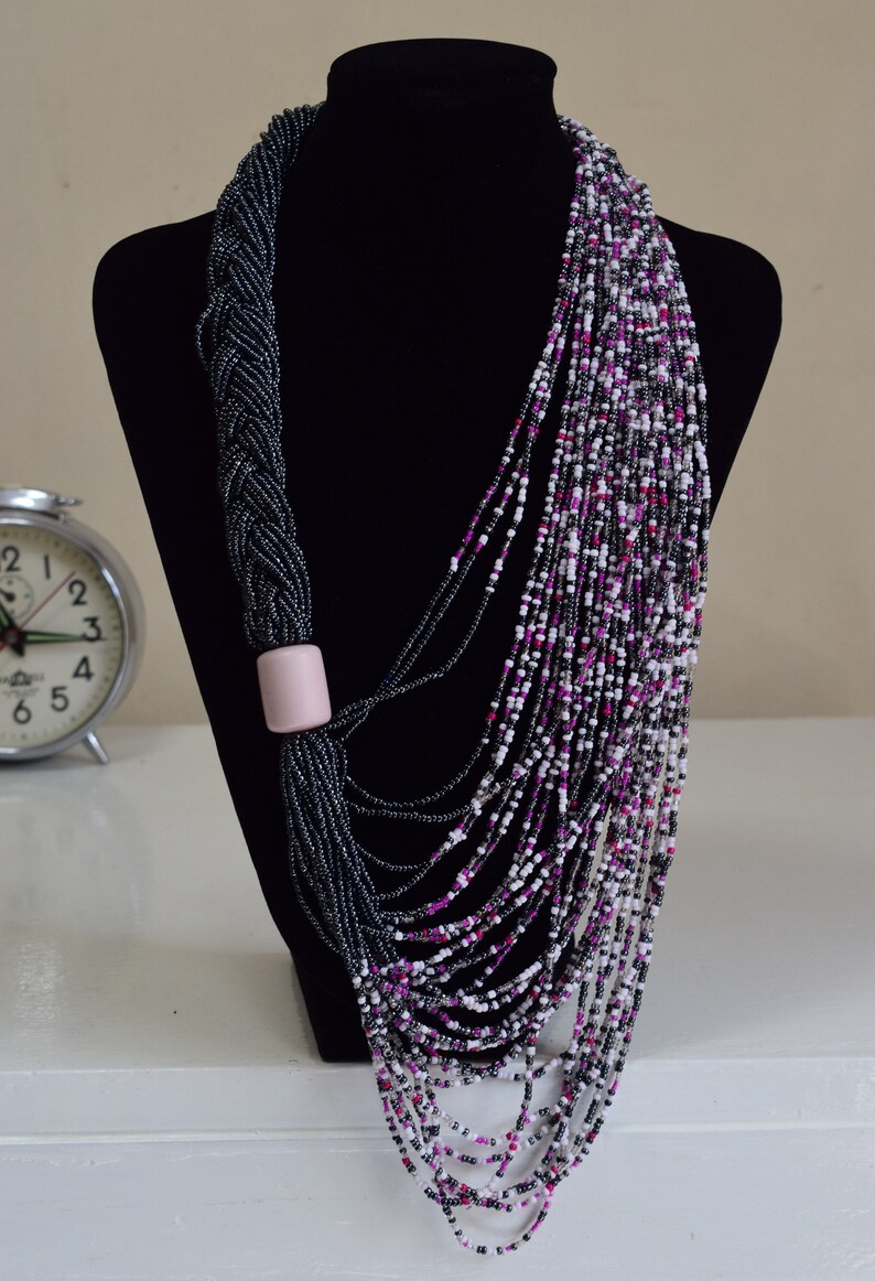 May include: A long, multi-strand necklace made of black, white, and pink beads. The necklace is draped over a black mannequin and features a pink bead accent.