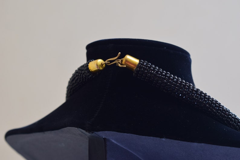 K&ouml;nnte beinhalten: Eine schwarze Perlenkette mit einem goldenen Verschluss. Die Kette wird auf einer schwarzen Samt-B&uuml;ste pr&auml;sentiert.