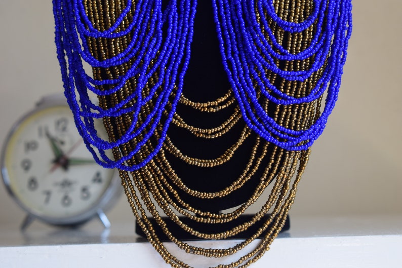 May include: A long, layered necklace made of blue and gold beads. The necklace is draped over a black mannequin.