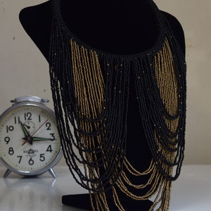 K&ouml;nnte beinhalten: Eine lange schwarze und goldene Perlenkette mit mehreren Perlenstr&auml;ngen, die von einem Choker-Stil nach unten fallen.