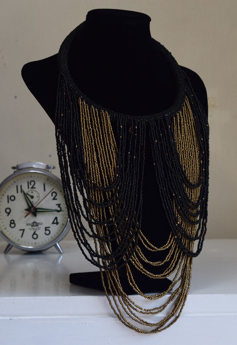 K&ouml;nnte beinhalten: Eine lange schwarze und goldene Perlenkette mit mehreren Perlenstr&auml;ngen, die von einem Choker-Stil nach unten fallen.