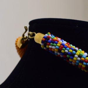 May include: A colorful beaded necklace with a gold-colored clasp and a small, orange pendant. The necklace is displayed on a black mannequin. The beads are a mix of blue, red, yellow, white, and black.