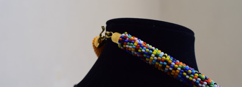 May include: A colorful beaded necklace with a gold-colored clasp and a small, orange pendant. The necklace is displayed on a black mannequin. The beads are a mix of blue, red, yellow, white, and black.