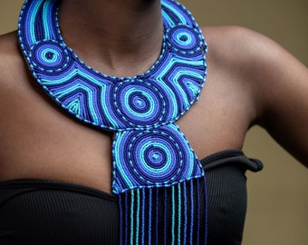 May include: A blue and turquoise beaded necklace with a large, circular pendant. The necklace is made of small, round beads that are strung together in a geometric pattern. The pendant is also made of beads and has a circular shape with a smaller circle in the center.
