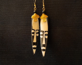 May include: A pair of black and white earrings with a tribal mask design. The earrings are made of wood and have gold accents.