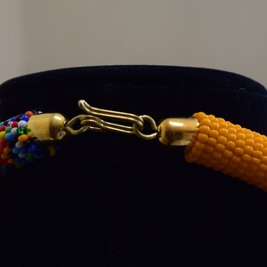May include: Close-up of a beaded necklace with a gold-colored clasp. The necklace features a section of colorful beads and a section of orange beads. The clasp is connected to gold-colored end caps.