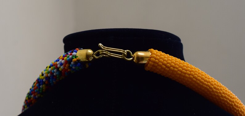 May include: Close-up of a beaded necklace with a gold-colored clasp. The necklace features a section of colorful beads and a section of orange beads. The clasp is connected to gold-colored end caps.