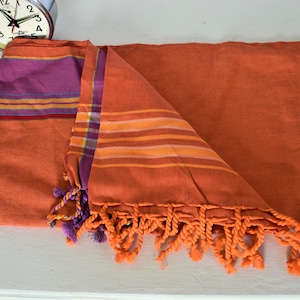 Könnte beinhalten: Orangefarbenes und violettes gestreiftes Strandtuch mit Quasten. Das Handtuch ist in der Hälfte gefaltet und hat eine gewebte Textur.